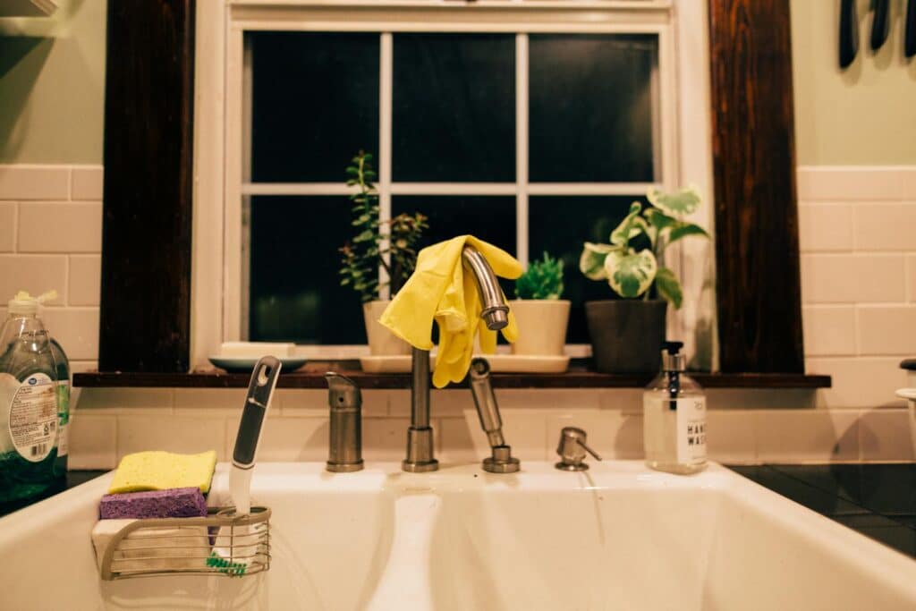 Warm lit kitchen sink at night with gloves, plants, and cleaning supplies.