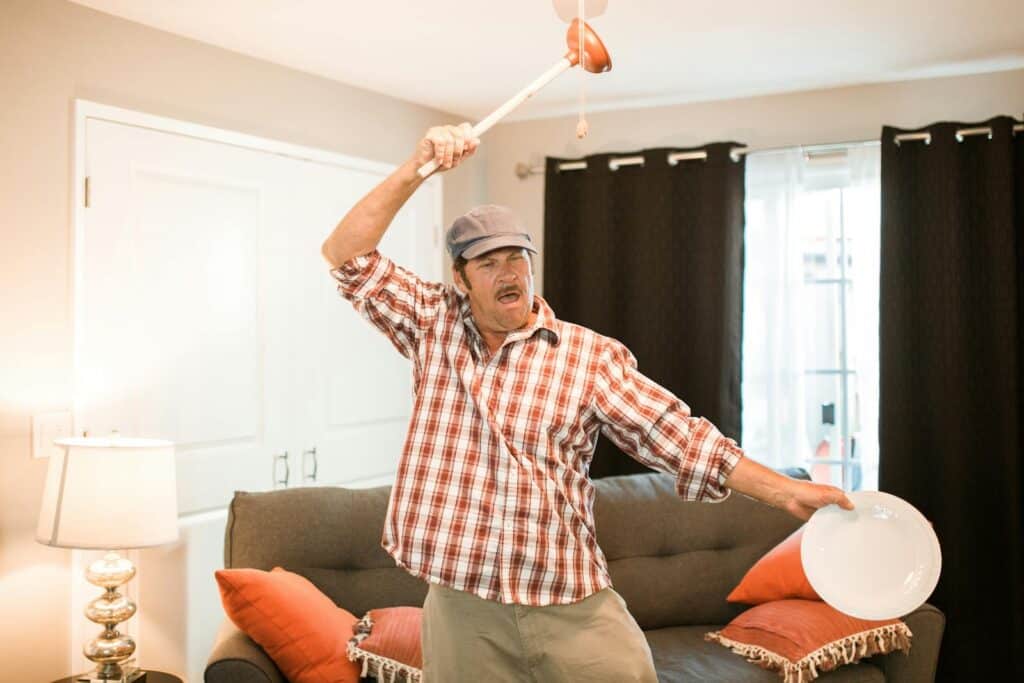 A cheerful man in a plaid shirt holding a plunger and plate in a lively manner inside a cozy living room.